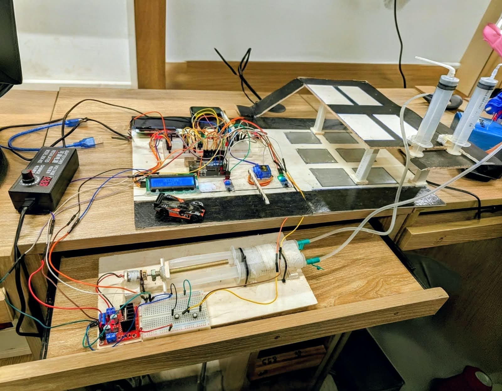 A model car park on a desk: an Arduino, IR sensors, a servo gate, an LCD reading Slot Left 2, and a syringe pneumatic lift driven by a motor and lead screw.