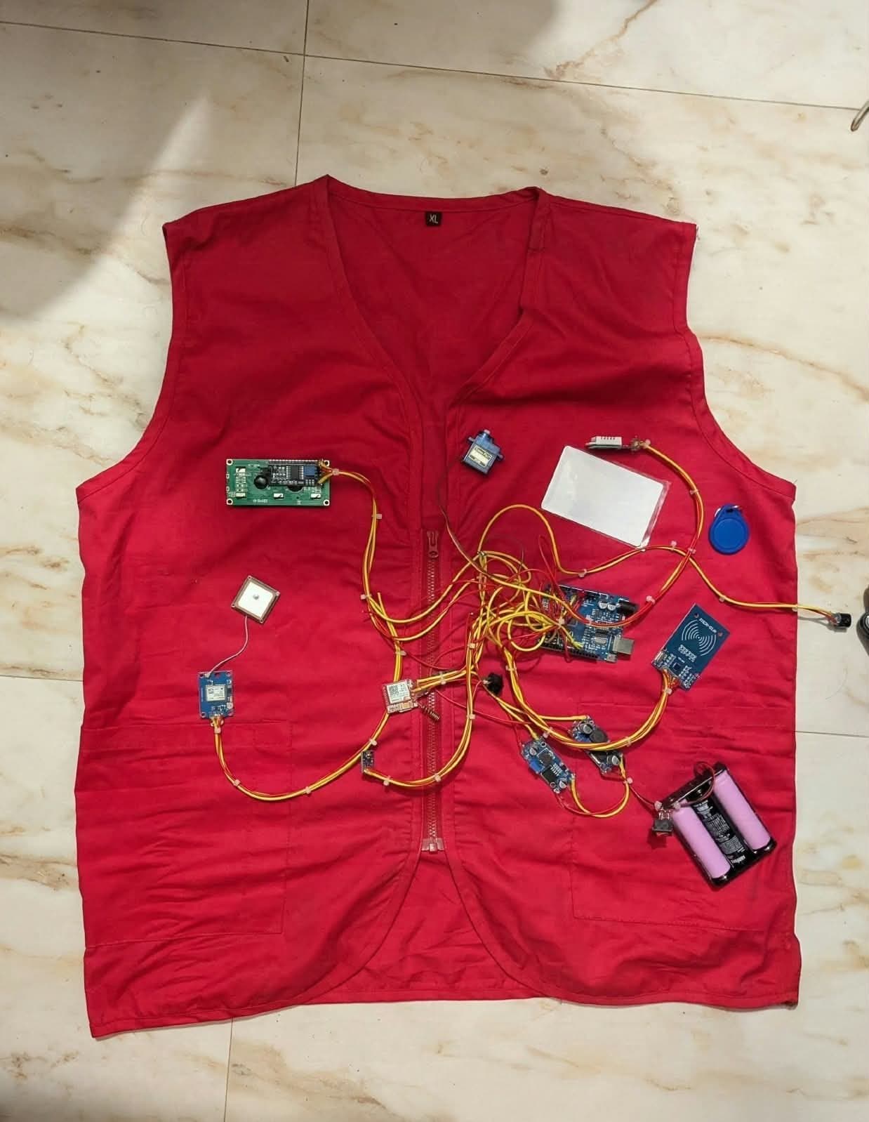 A red work vest laid flat with an Arduino, RFID reader, GPS and GSM modules, a servo, a display and a battery pack wired across it.