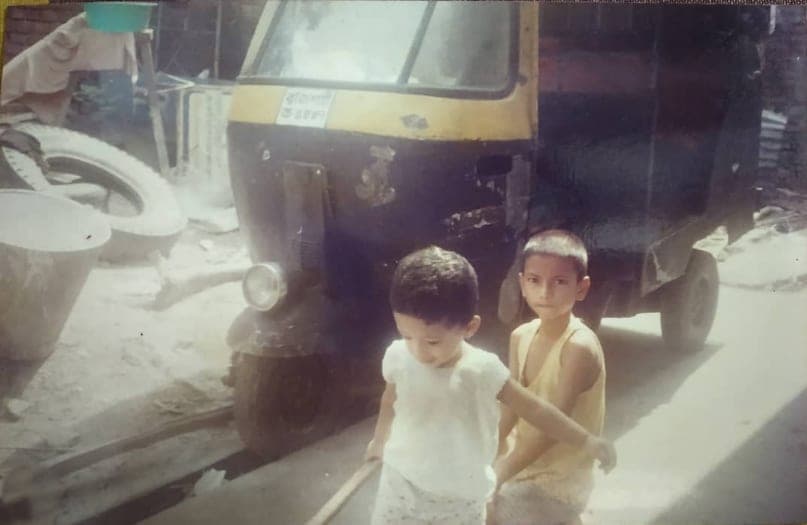 Two small children on a Rajshahi street, an auto-rickshaw parked behind them.